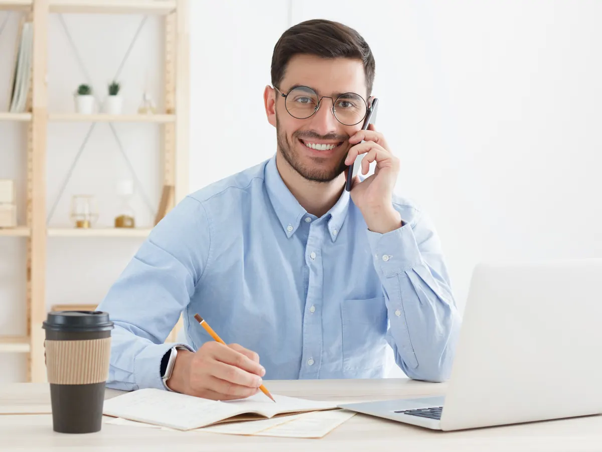 A man on a phone call, smiling and taking notes.